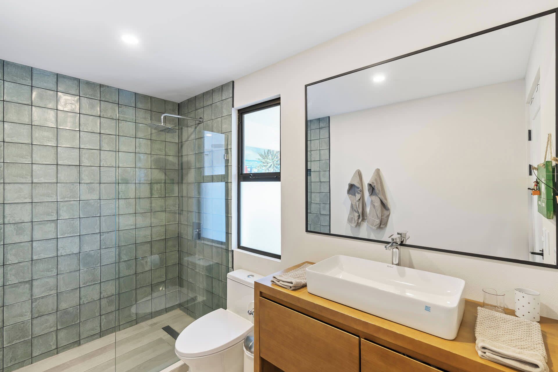 Un baño moderno con cabina de ducha de vidrio, inodoro blanco, tocador de madera con un espejo grande y lavamanos blanco.
