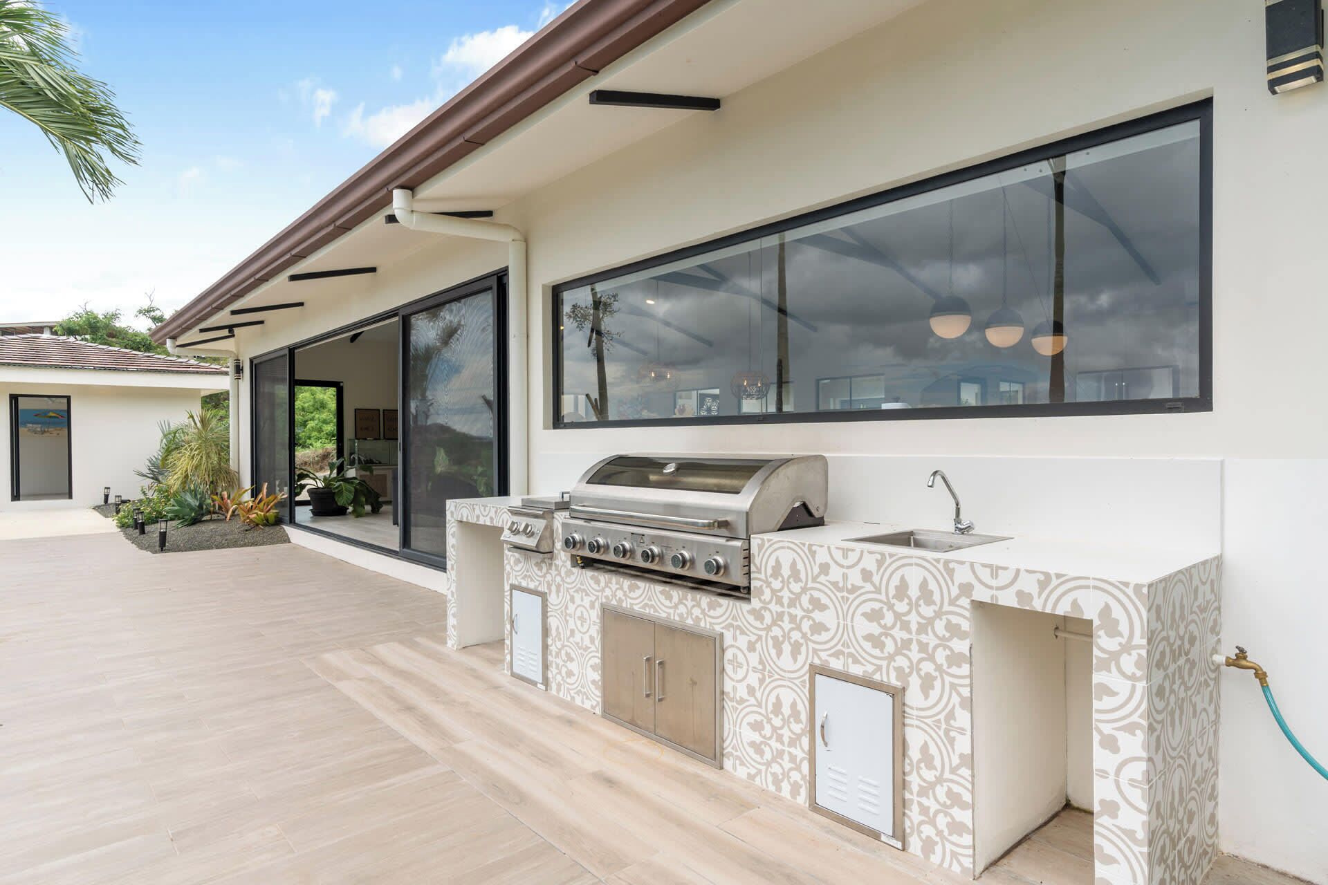 An outdoor kitchen with a grill, sink, and storage cabinets, adjacent to a large window overlooking a garden.