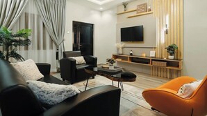 A modern living room with a black sofa, an orange armchair, a black coffee table, and a TV mounted on the wall. - Kashco Apartments (Abuja)