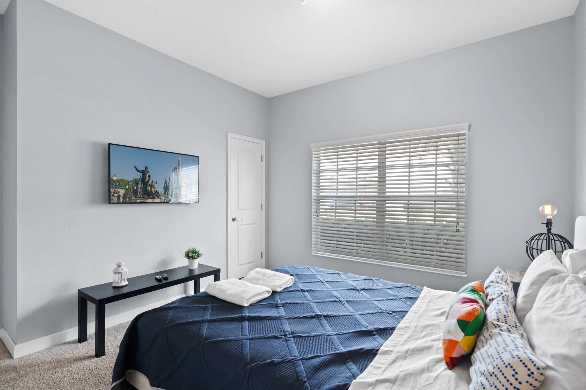 A bedroom with a bed, a nightstand, a window with blinds, a door, and a wall-mounted picture.
