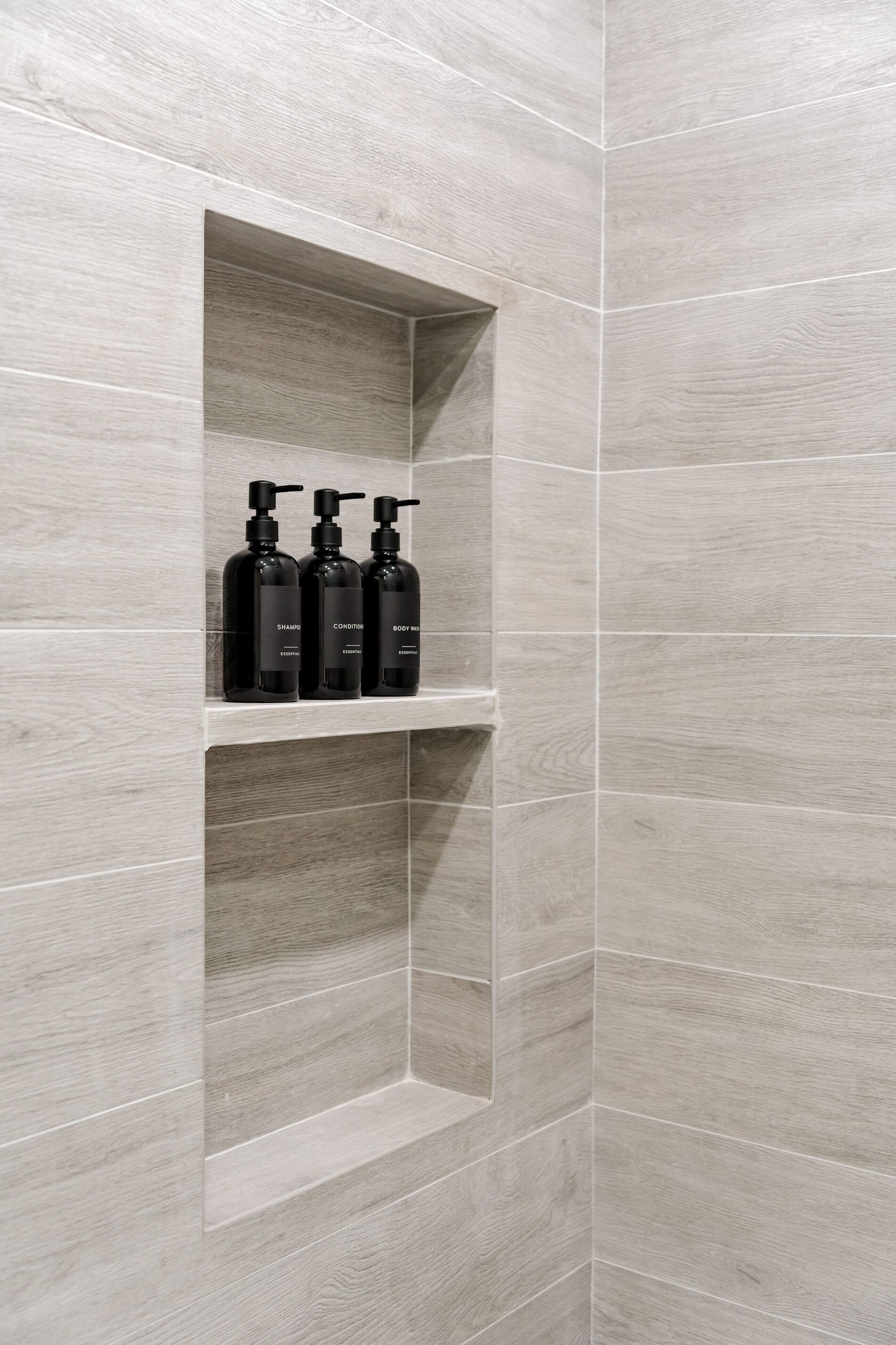 A bathroom with grey tiles and a shelf holding three black bottles of shampoo, conditioner, and body wash.