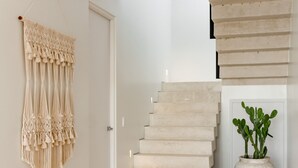 Eine Treppe mit beigen Stufen führt zu einer Zwischenebene, dort ist ein Deckenventilator, ein Wandbehang aus Makramee und ein Topfpflanze zu sehen.