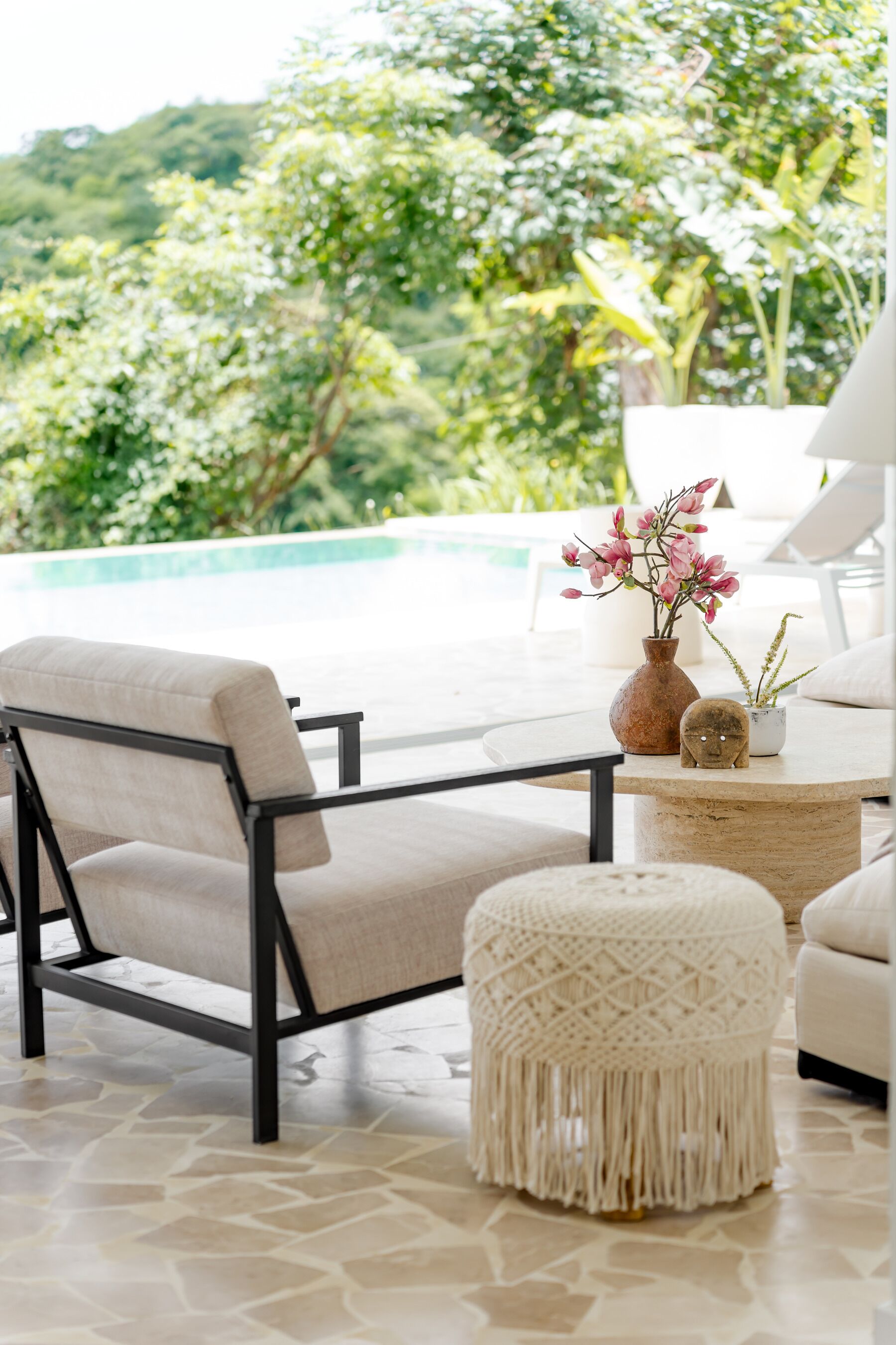 A sunlit outdoor seating area with a pool view, a white cushioned chair, a small round ottoman, and a vase with pink flowers on a table.