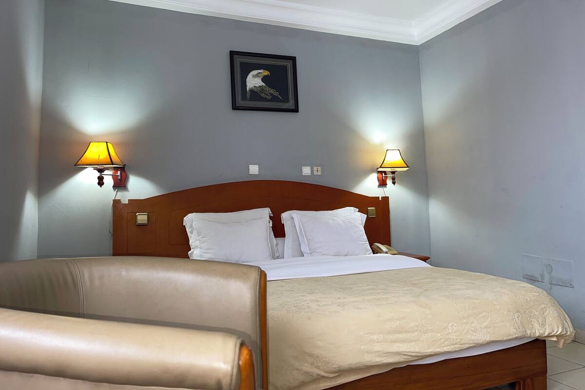 Une chambre d’hôtel avec une tête de lit en bois, deux lampes de chevet, un tableau encadré au mur et un fauteuil beige.