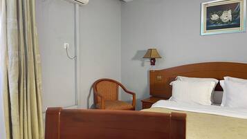 Una camera d'albergo con un letto di legno, cuscini bianchi, una sedia di legno, un comodino, una lampada e un quadro appeso al muro.