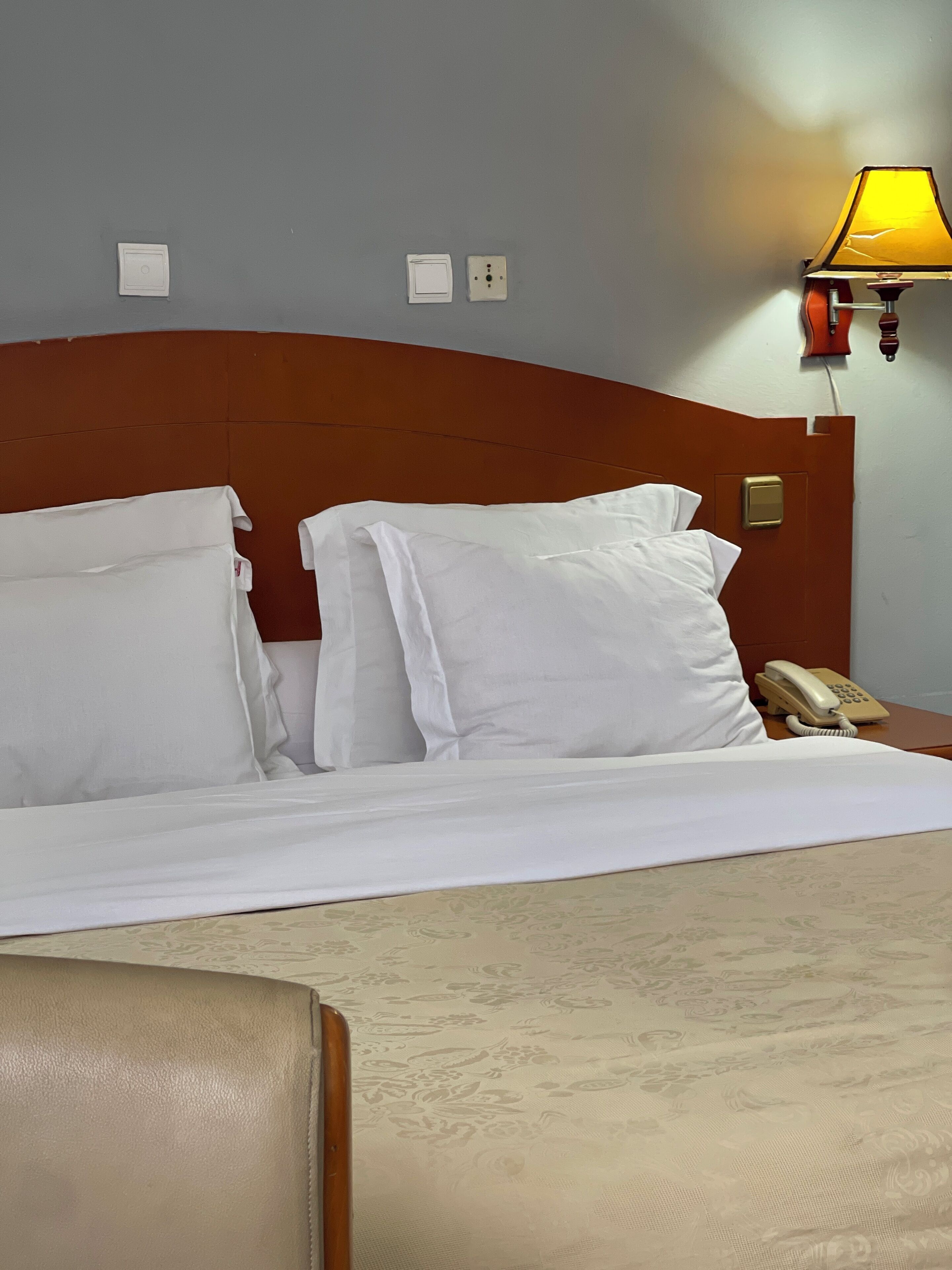 A hotel room with a bed, pillows, a bedside table with a lamp, and a telephone.