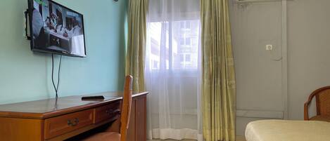 A hotel room with a wooden desk, a television mounted on the wall, a bed with a white bedspread, and a window with sheer curtains.