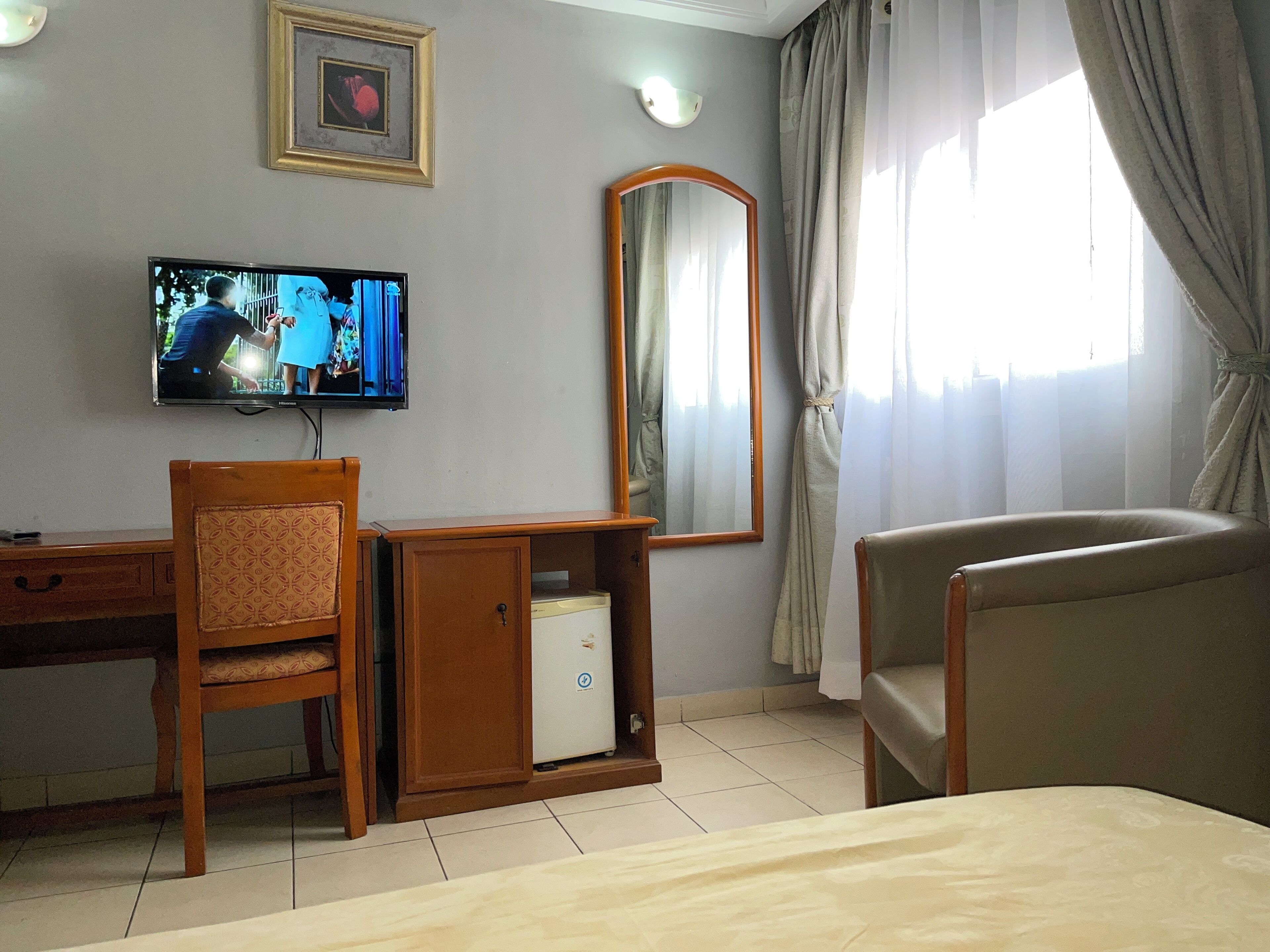 A hotel room with a bed, a desk with a chair, a television, a mirror, and a window with curtains.