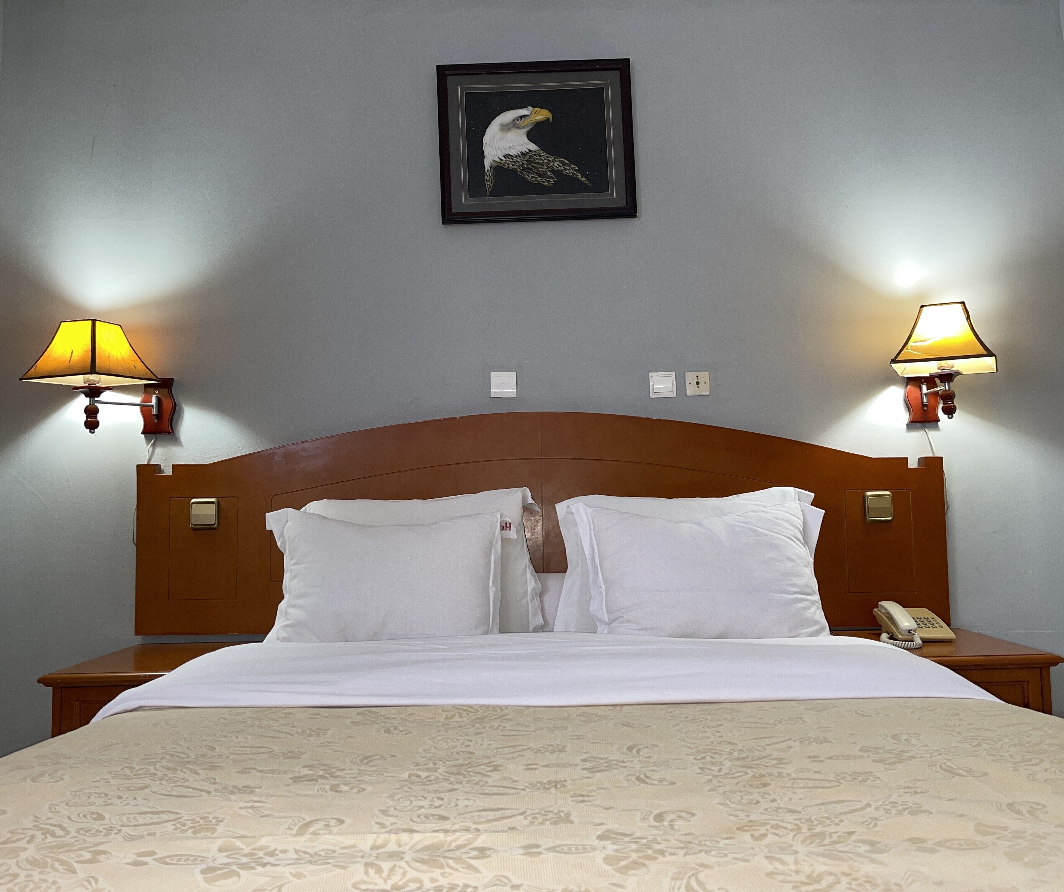 A hotel room with a wooden headboard, two bedside lamps, a framed picture above the bed, and a telephone on the nightstand.
