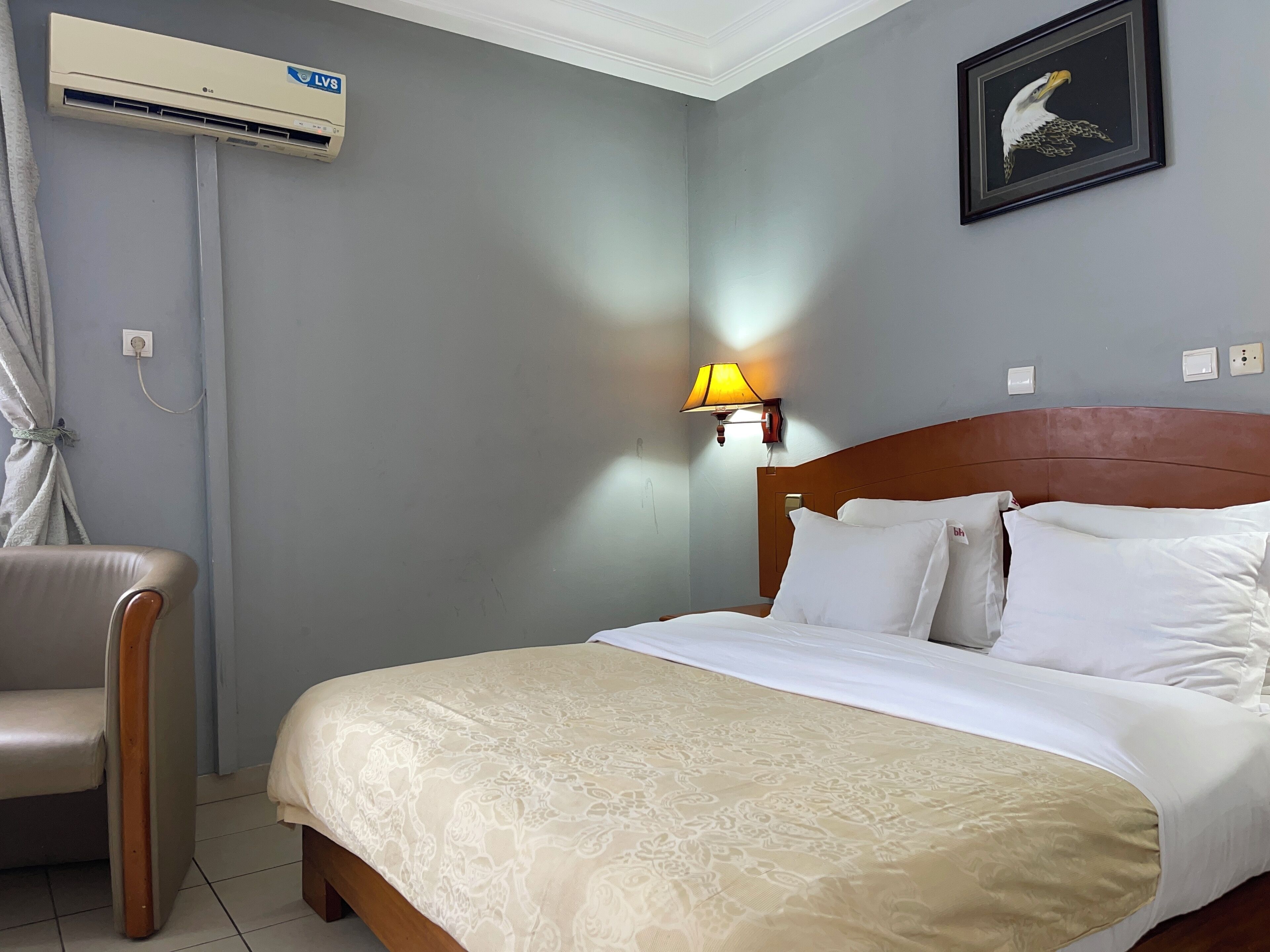 Une chambre d’hôtel avec un lit, une chaise, un appareil de climatisation, une lampe fixée au mur et un tableau encadré accroché au mur.