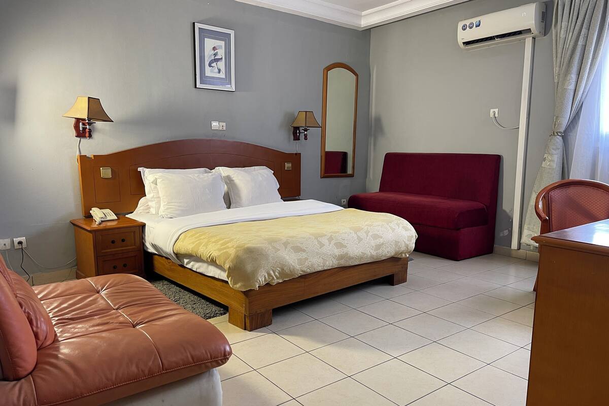 Une chambre d’hôtel avec un lit en bois, un fauteuil rouge, un canapé en cuir marron, une table de chevet avec une lampe et un appareil de climatisation.