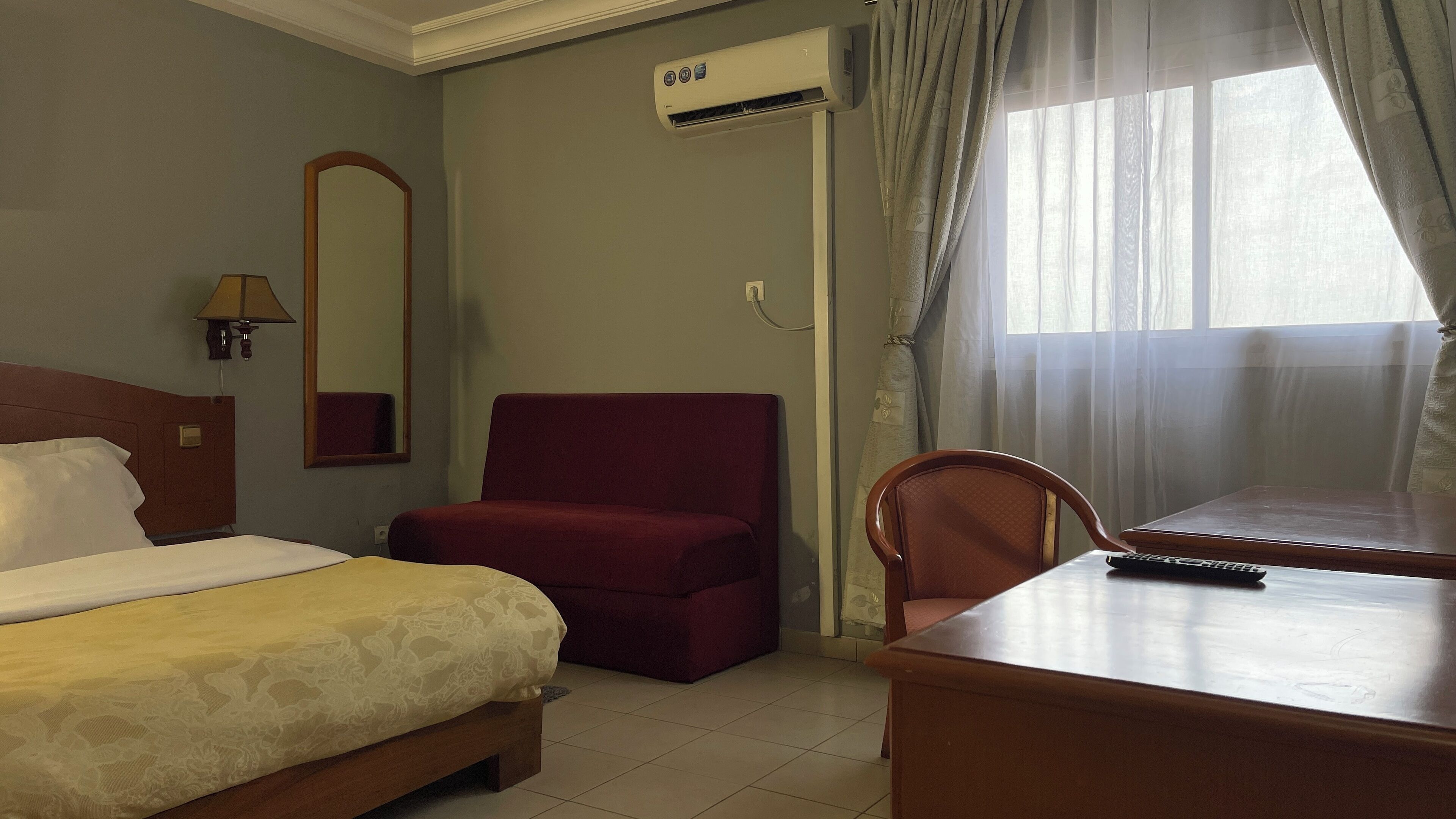 A hotel room with a bed, a red armchair, a wooden desk, and a window with curtains.