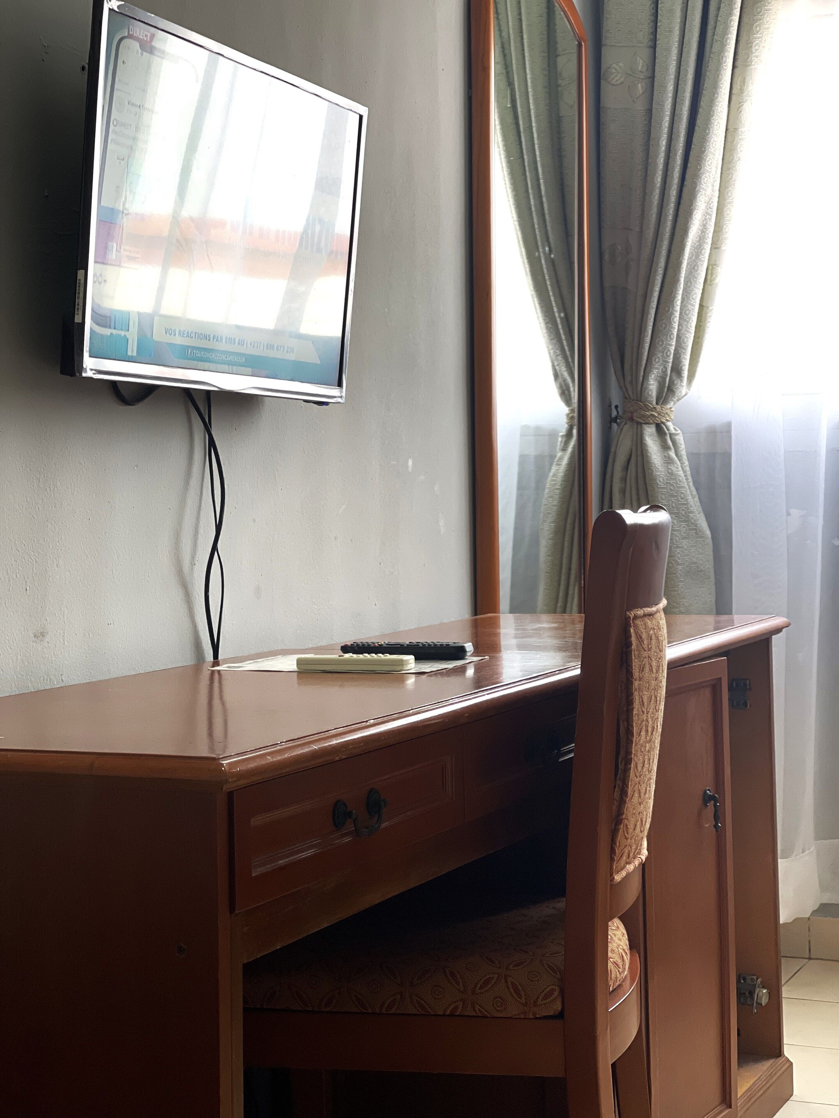 A wooden desk with a television mounted on the wall, a remote control, and a mirror.