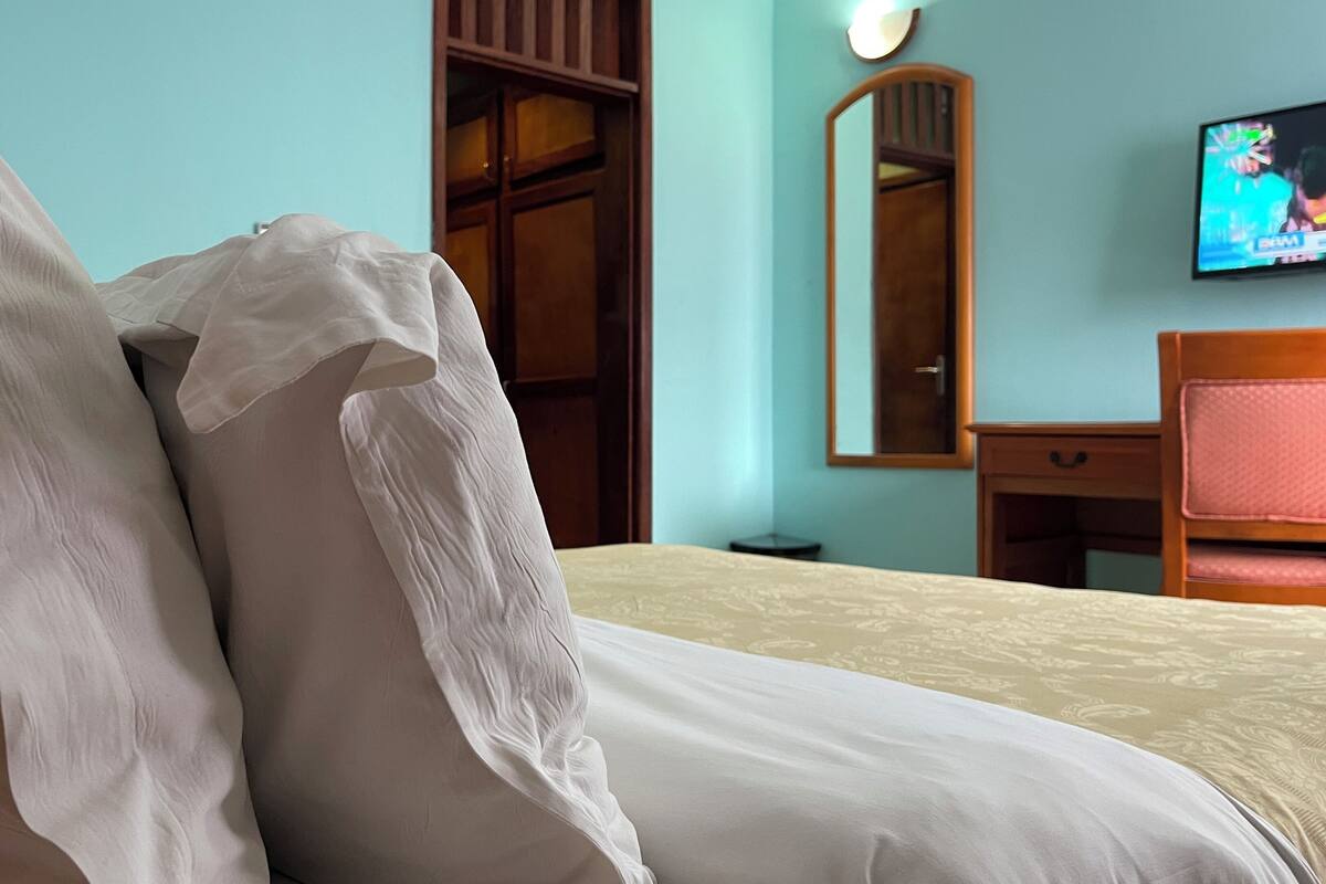 Un lit avec une literie blanche et beige, une armoire en bois, un miroir et un téléviseur fixé au mur.