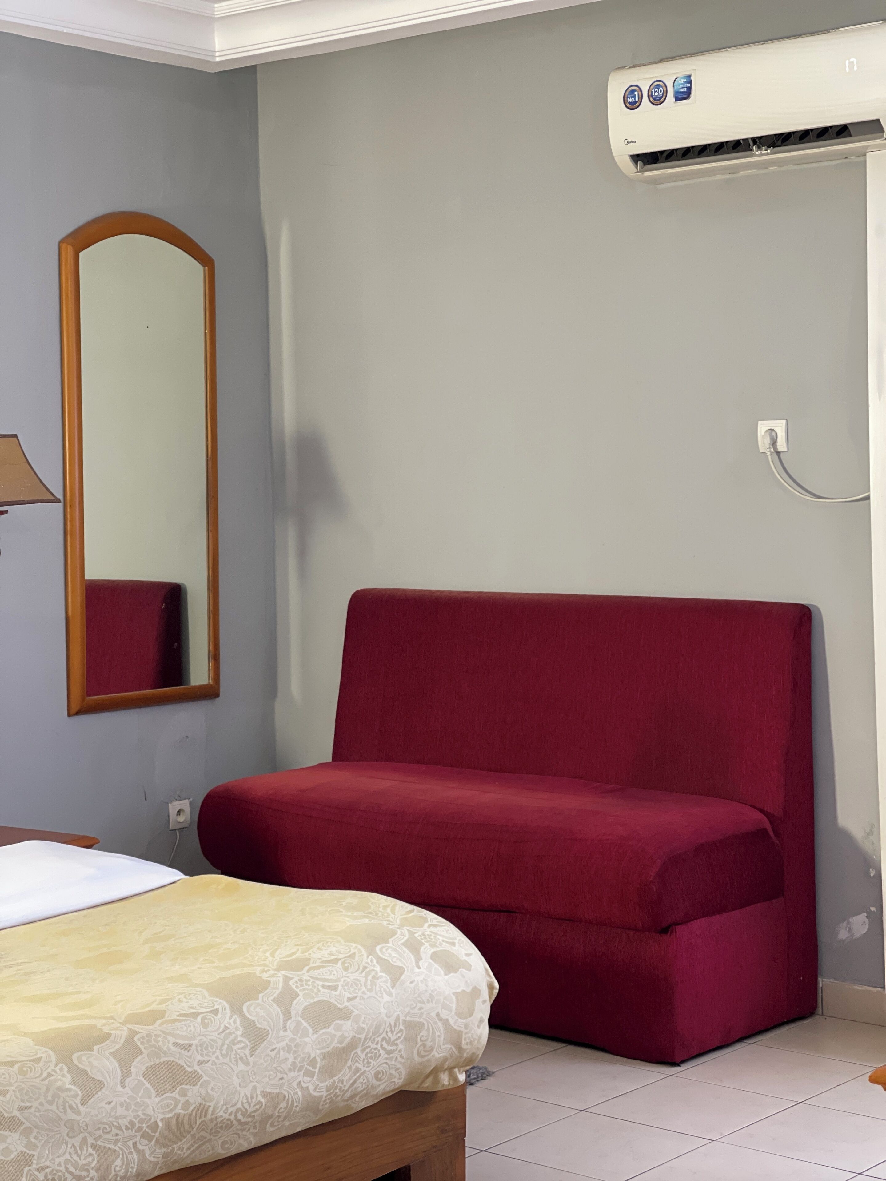 Una habitación con un sofá rojo, una cama con colcha estampada, un espejo y un equipo de aire acondicionado.