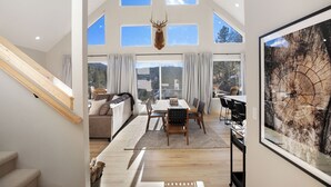 A spacious room with a high ceiling, large windows, a dining area with a wooden table and chairs, a mounted deer head, and a large framed tree trunk artwork. - 3BR Divide Cabin: Starlink, Mtn Views, Hiking Access! Family Friendly - Sleeps 8 (Divide)