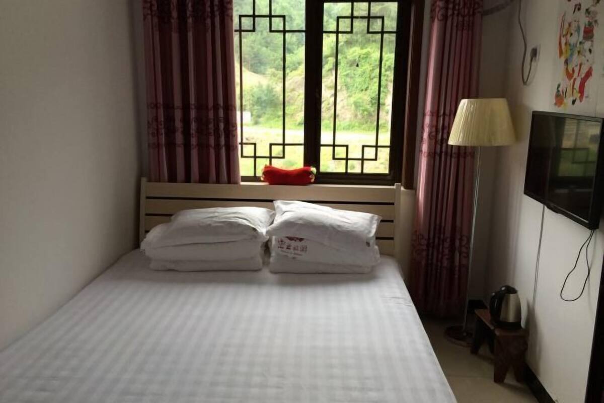 A bed with pillows, a window with curtains, a lamp, and a television on the wall.
