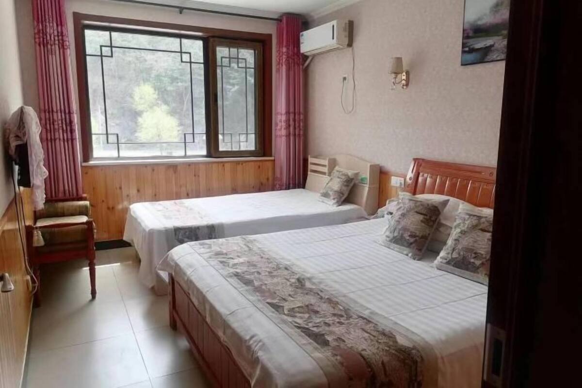 A room with two beds, a wooden chair, a window with curtains, an air conditioning unit, and a wall-mounted lamp.