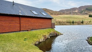 Una cabaña de troncos con techo inclinado y tragaluces, junto a un lago tranquilo con orilla de césped.