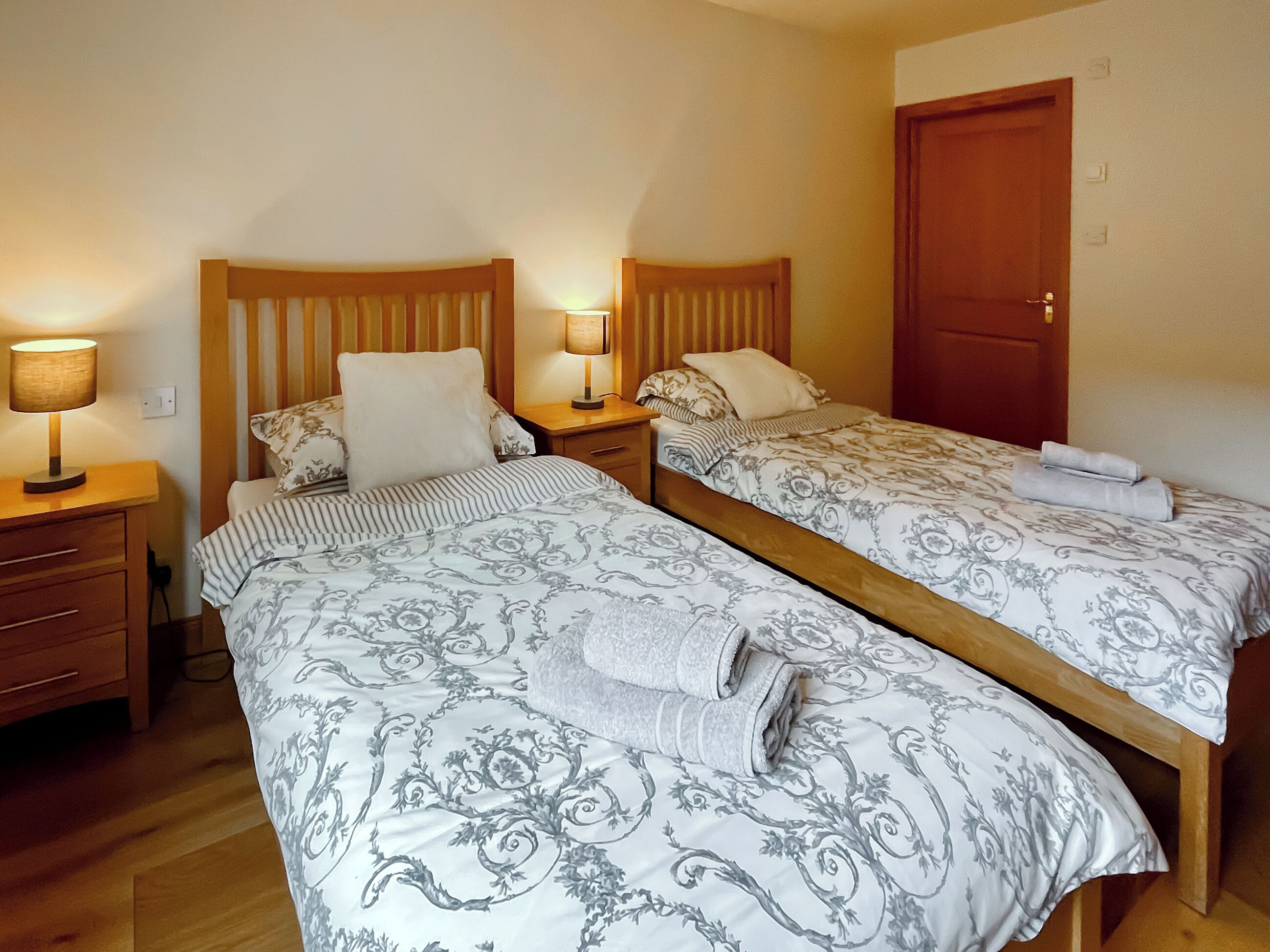 Een tweepersoonskamer met twee eenpersoonsbedden, elk voorzien van een houten hoofdbord, een nachtkastje met een lamp en een opgevouwen handdoek op het bed.