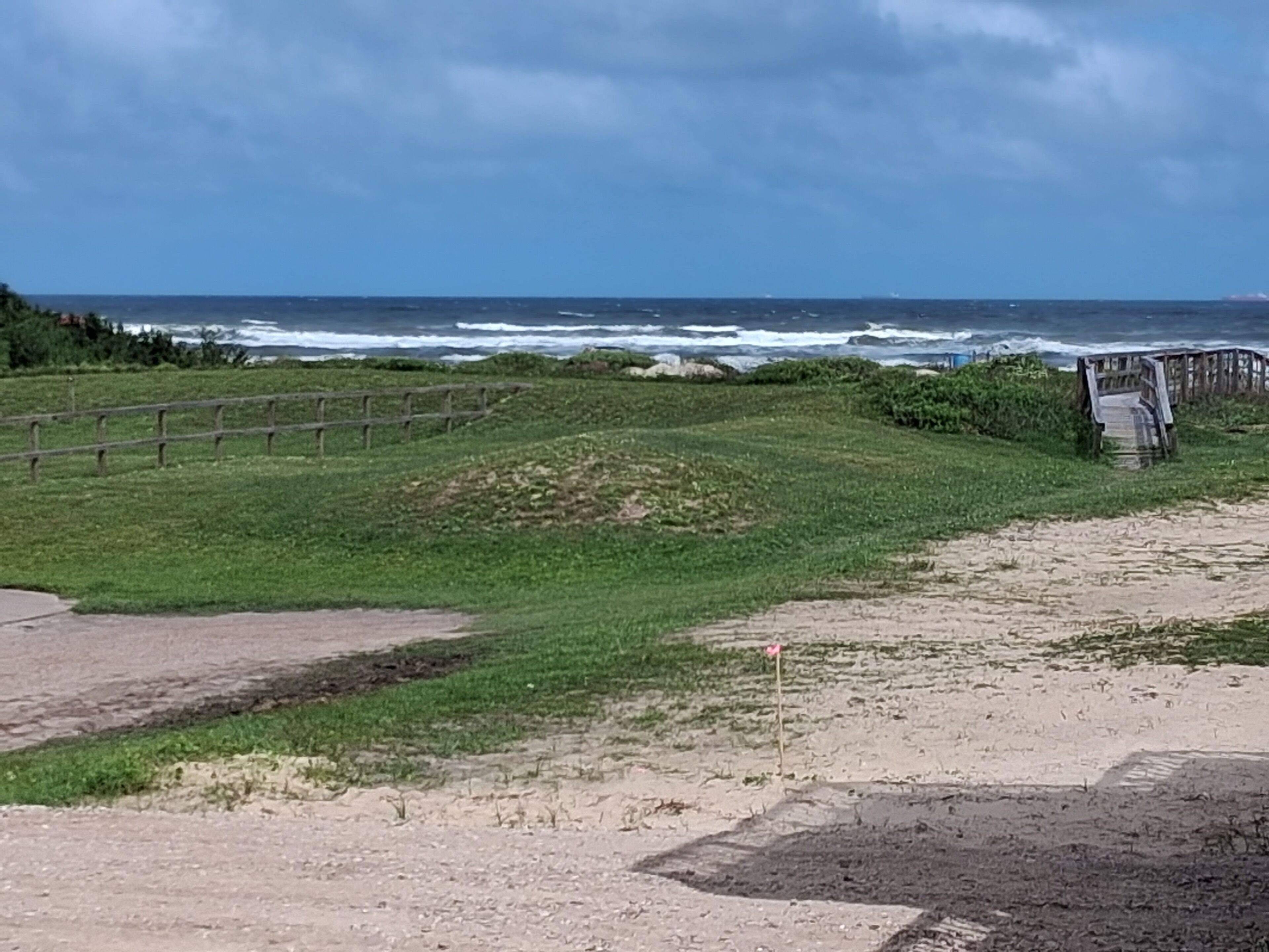 A coastal landscape with a grassy area, a wooden fence, and a sandy path leading to a beach.