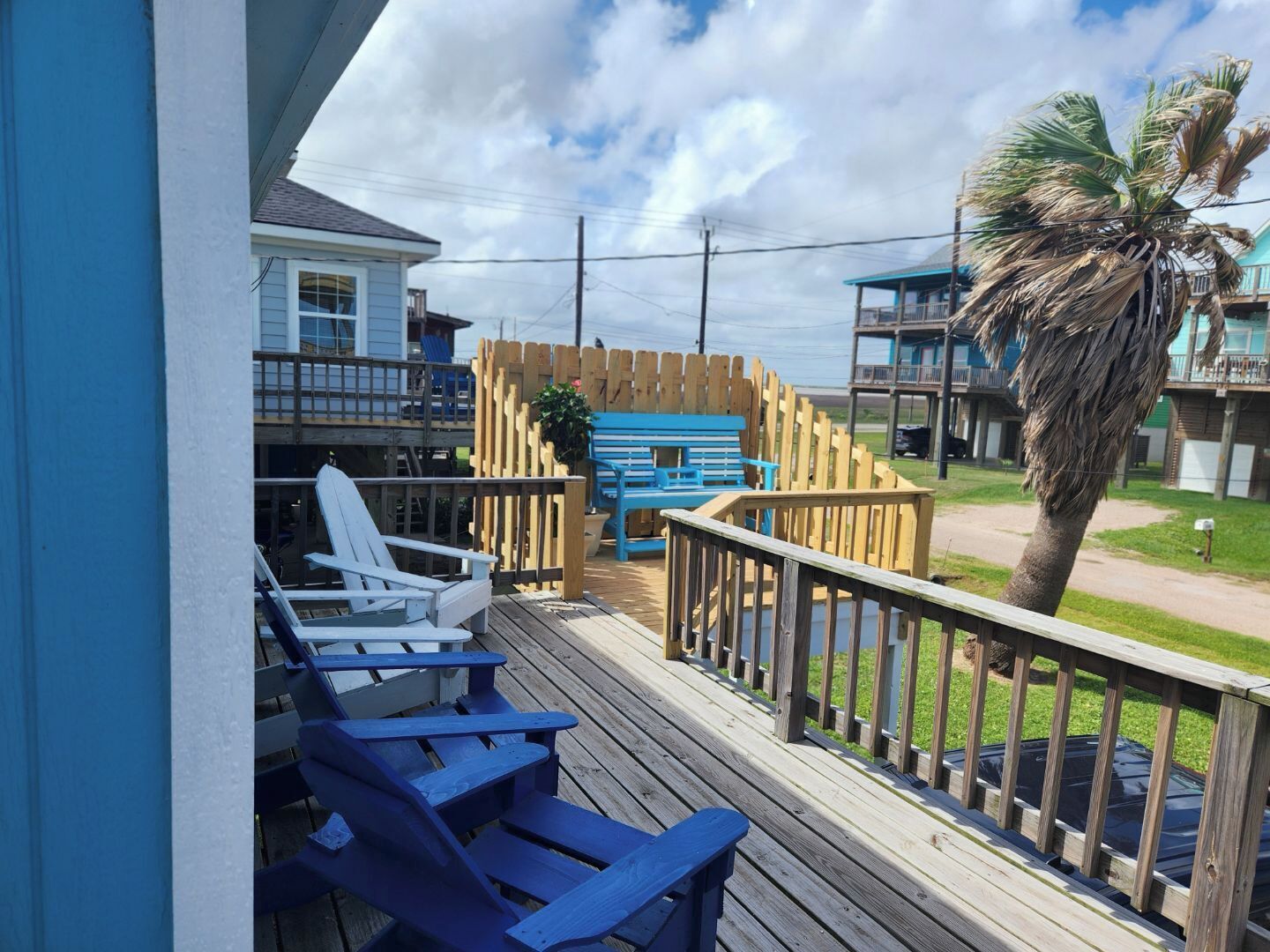 A wooden deck with blue Adirondack chairs, a wooden fence, and a view of a beach and buildings.