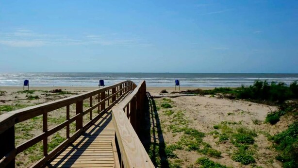 Ein Holzsteg führt zu einem Strand mit zwei blauen Stühlen und freiem Blick auf das Meer.