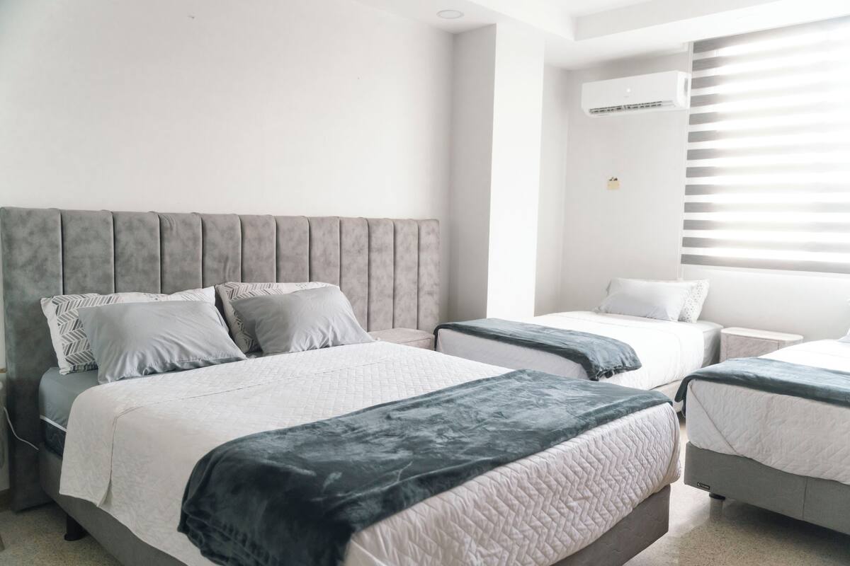 A hotel room with two beds, a grey headboard, and a window with blinds.