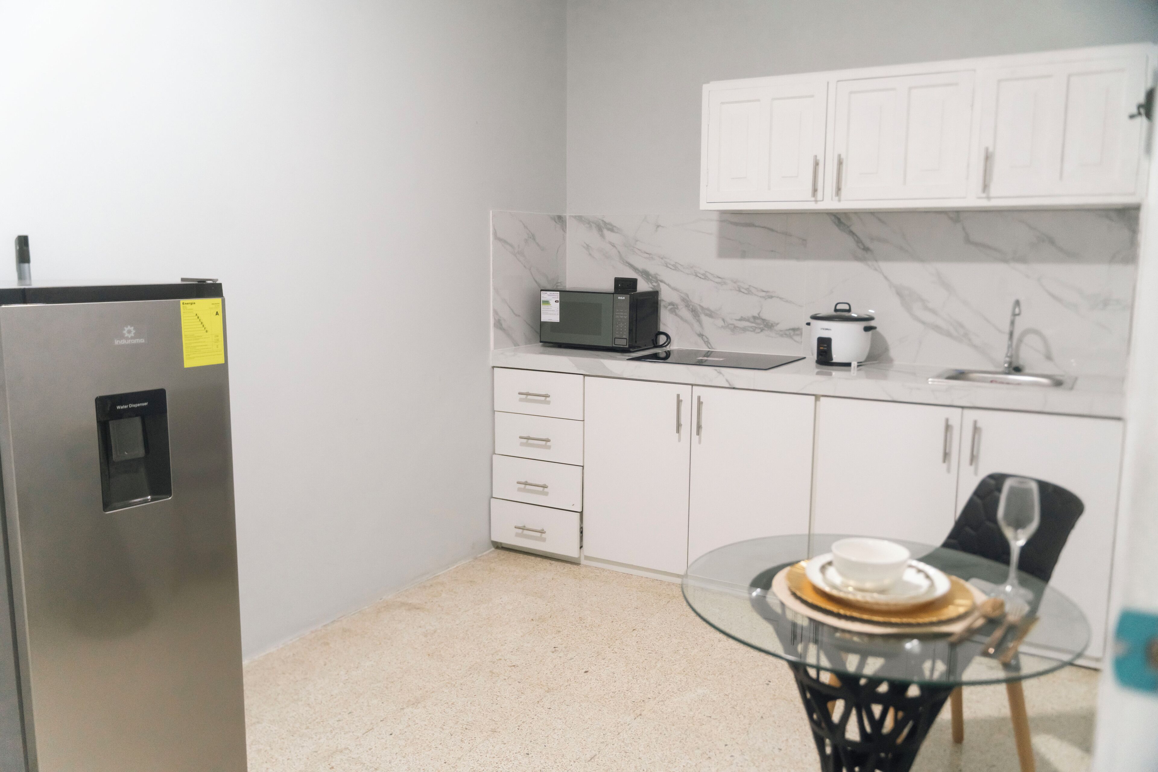 A compact kitchen with a refrigerator, microwave, and stove, white cabinets, and a glass table with a cup and saucer.