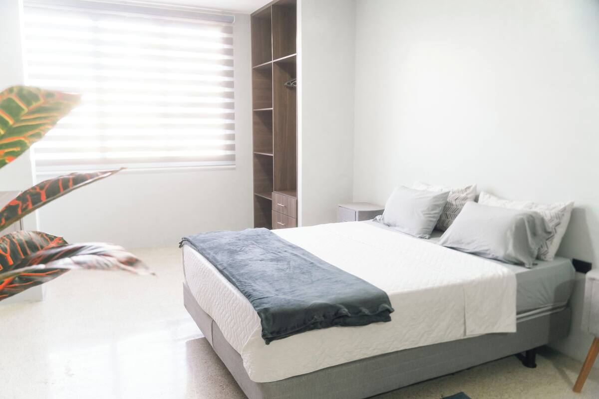 A bedroom with a bed, a nightstand, a wardrobe, and a large window with blinds.