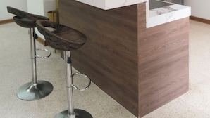 A modern kitchen with a marble countertop, a built-in island, two bar stools, a hanging light fixture, a refrigerator, and a potted plant. - Guayacan Apart Hotel  (Guayaquil)