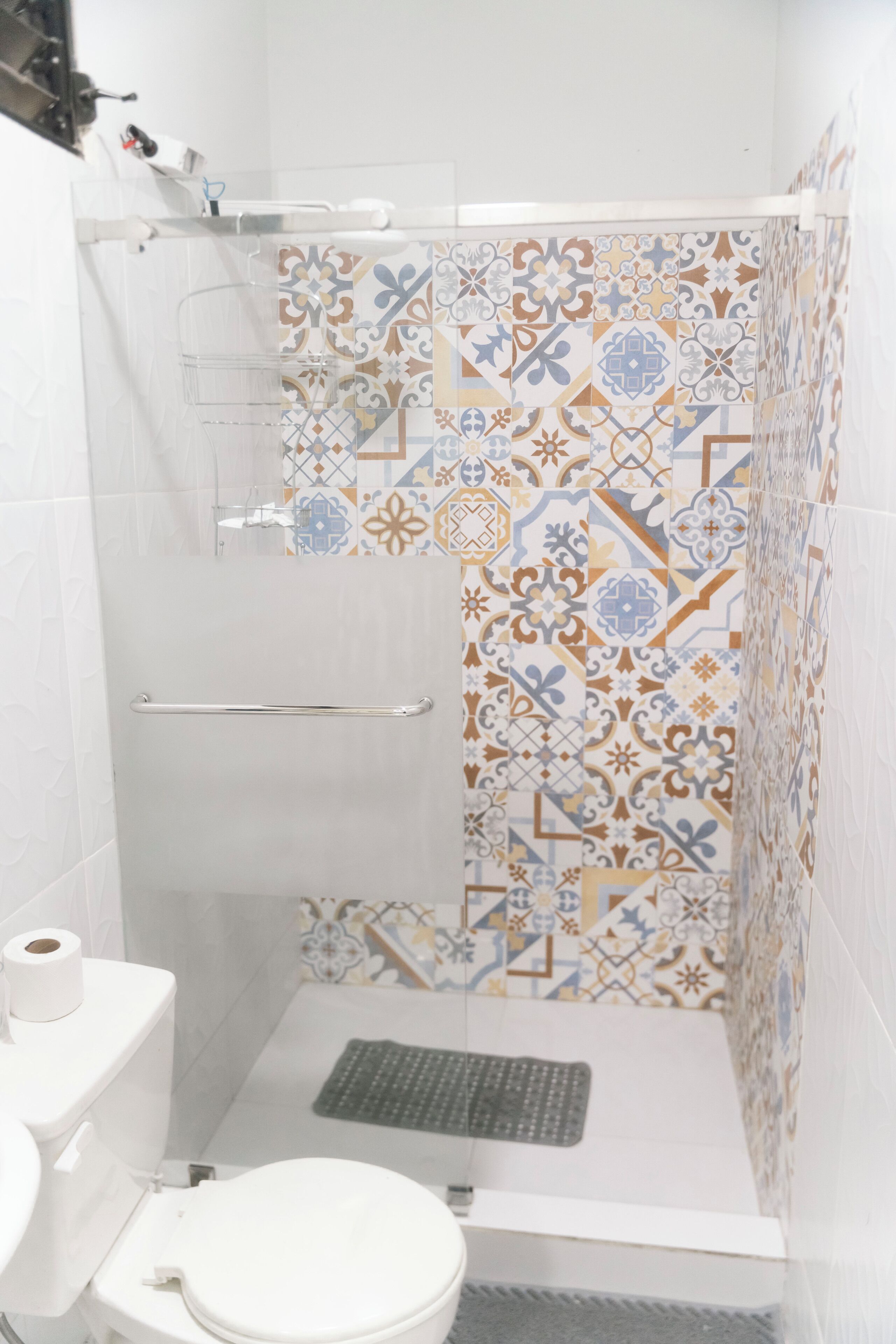 Une douche avec un mur recouvert de carreaux à motifs, une barre d’appui et des toilettes avec un rouleau de papier toilette.