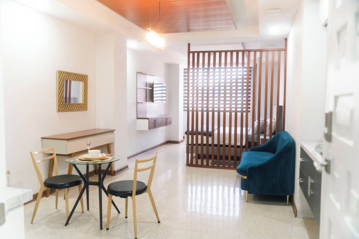 A modern hotel room with a dining area, a blue sofa, a wooden partition, and a small table with chairs.
