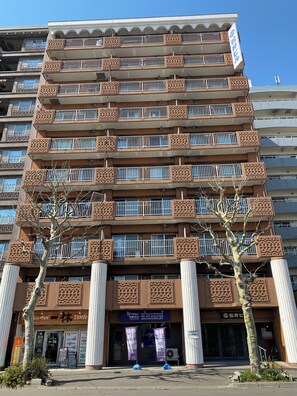Un immeuble à plusieurs étages doté de balcons, une devanture de magasin avec une enseigne, et un arbre devant.