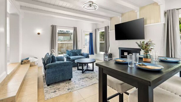 A modern living room with a sofa set, a dining table with plates and glasses, and a flat-screen TV mounted on the wall. - Anastasia Island Retreat (St. Augustine)