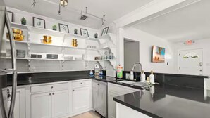 A modern kitchen with white cabinetry, black countertops, and a built-in island. There are shelves above the sink holding various kitchen items and a painting on the wall. - Anastasia Island Retreat (St. Augustine)