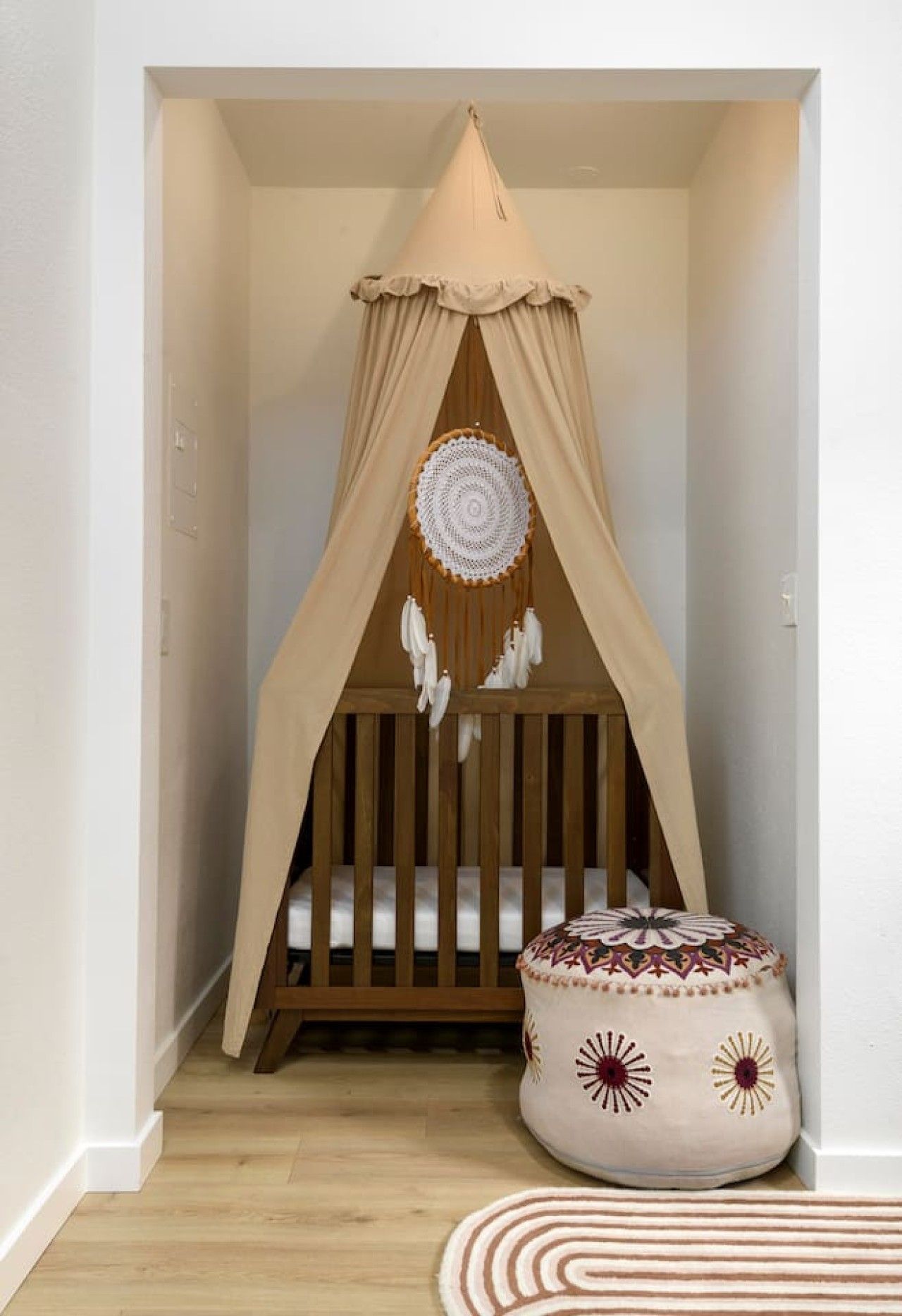 A cozy baby crib with a canopy, a decorative dreamcatcher, and a cushioned ottoman in a hallway.