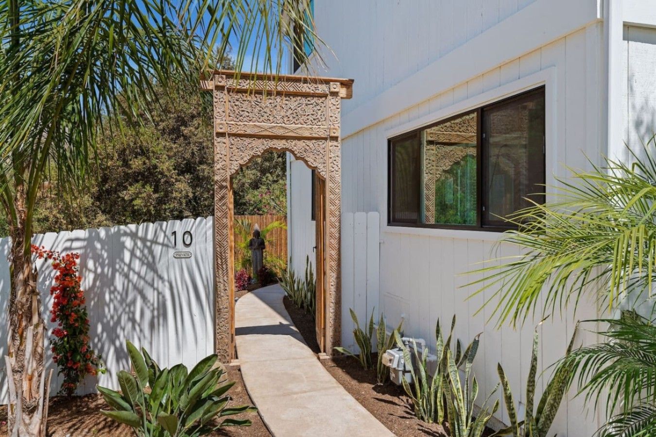 A pathway leading to a building with a decorative archway, a white fence, and various plants.