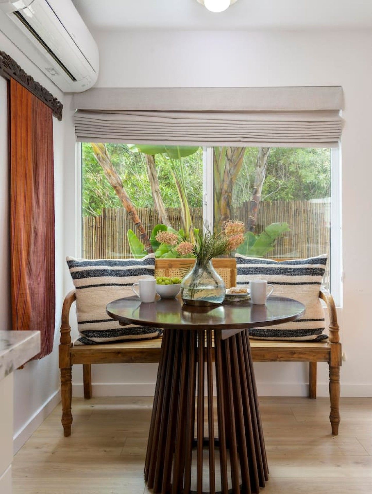 A room with a wooden bench, a round table, and a window with a view of greenery.