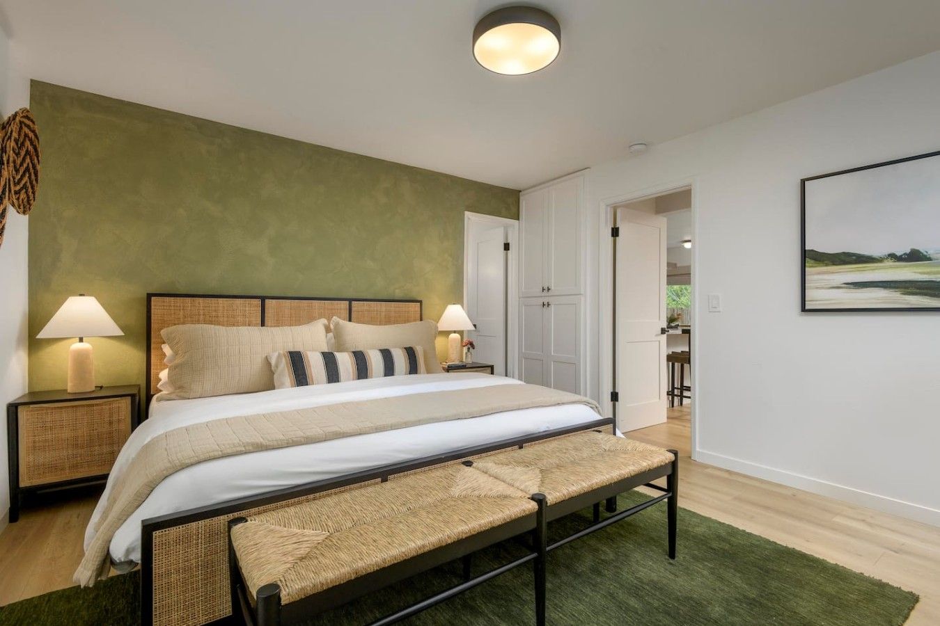 A bedroom with a bed, bedside lamps, a bench, a green wall, and a door leading to another room.