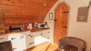 A cozy kitchen with wooden cabinets, a sink, and a microwave. There's a door with a sign, a chair, and a framed welcome sign on the wall. - Cabanau Bach (Caerphilly)