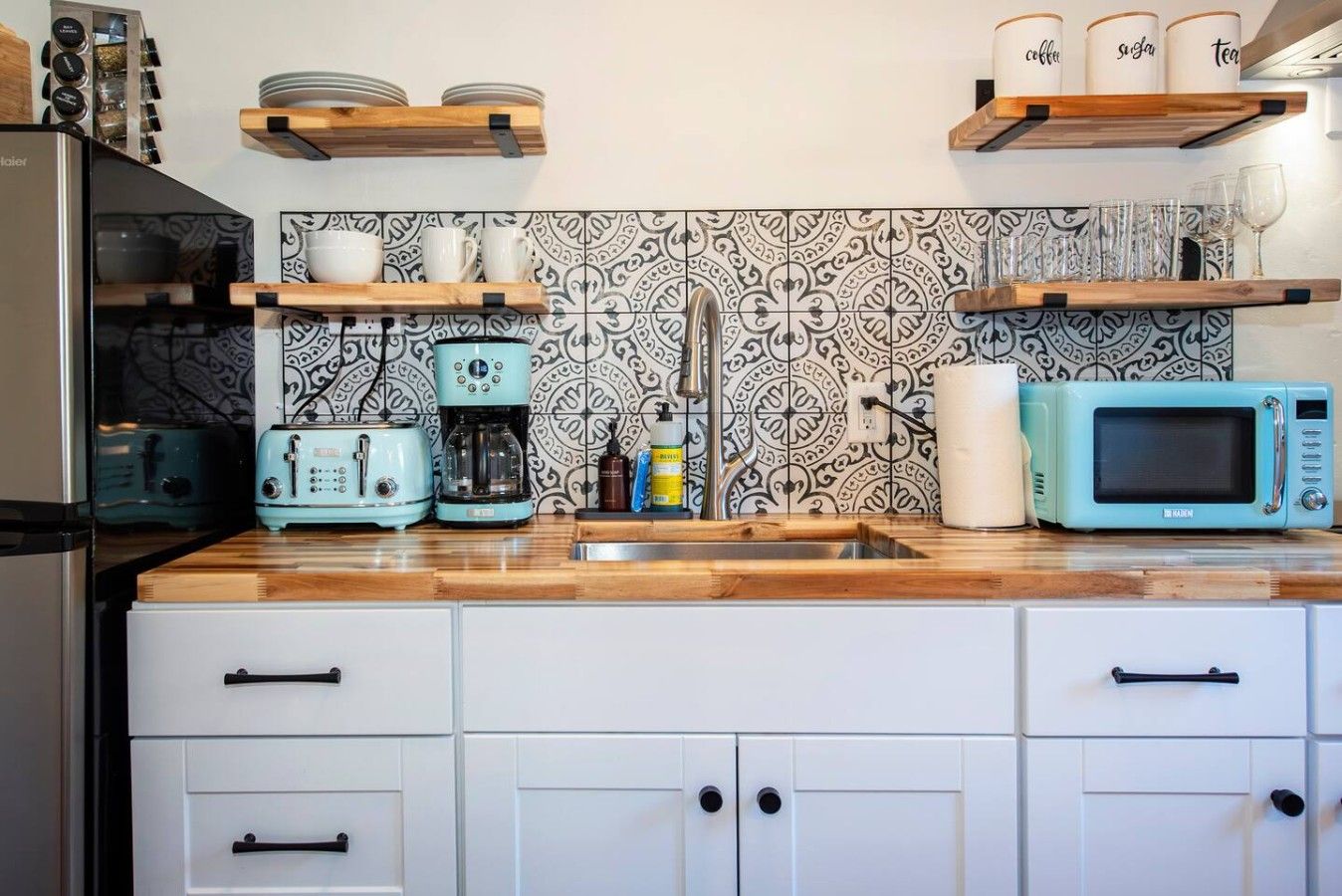 Una cucina con mobili bianchi, un piano di lavoro in legno, una macchina per il caffè, un tostapane, un forno a microonde e mensole con tazze e barattoli.