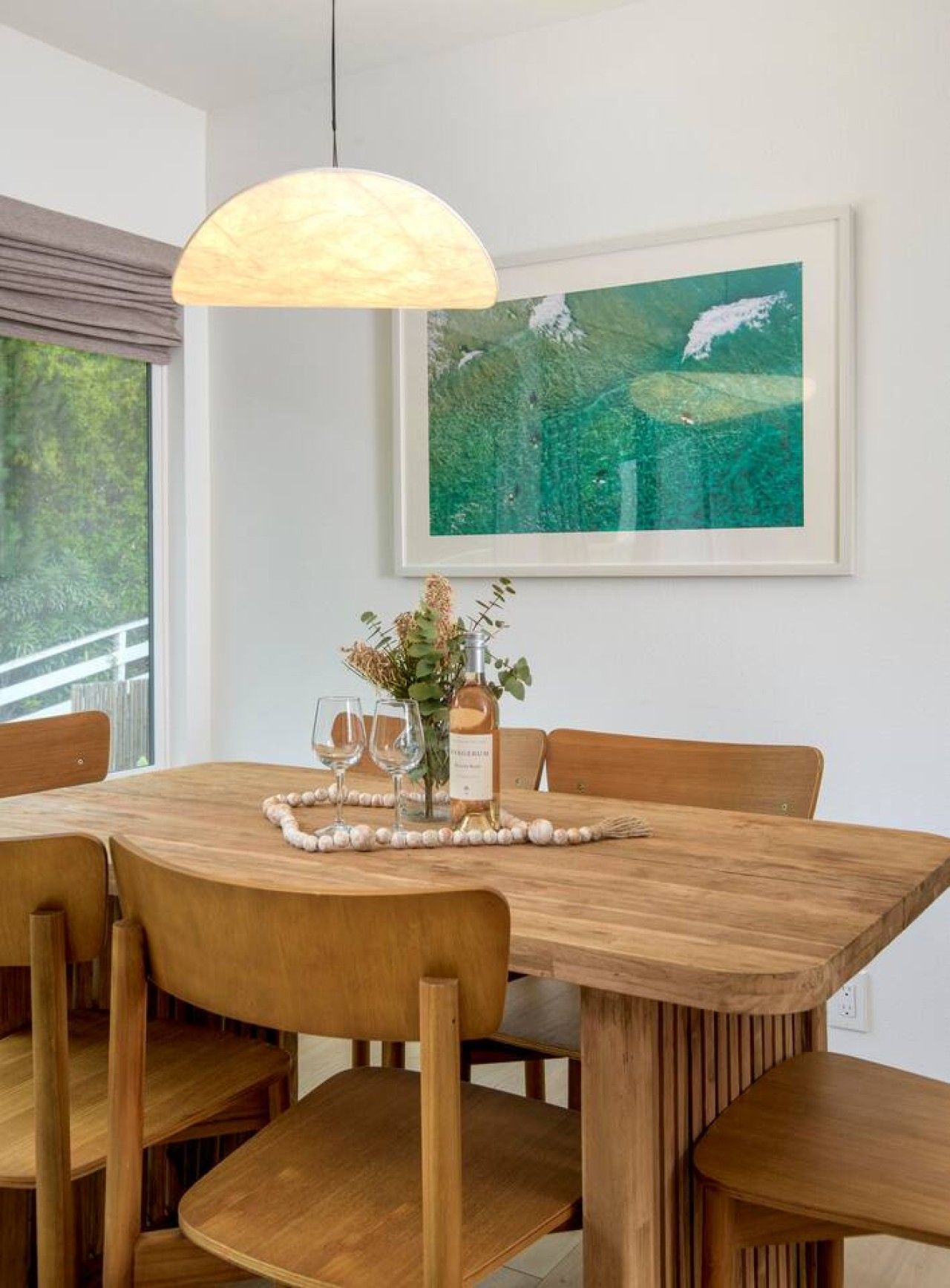 Een eetgedeelte met een houten tafel, stoelen, een hanglamp, een lijst met een schilderij en een vaas met bloemen.