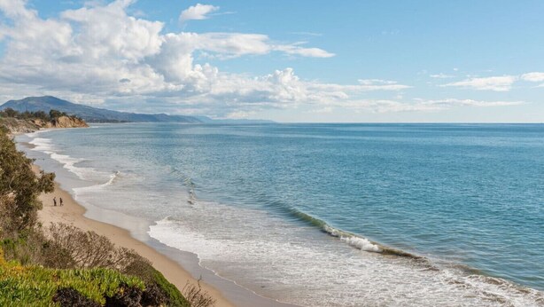 Ein Strand mit klarem, blauem Wasser, einem sandigen Ufer und Bergen im Hintergrund.