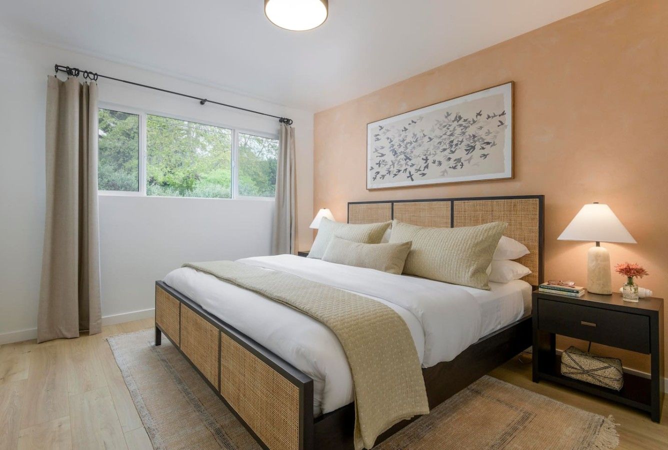 A bedroom with a bed, bedside lamps, a window with curtains, and a wall-mounted artwork.