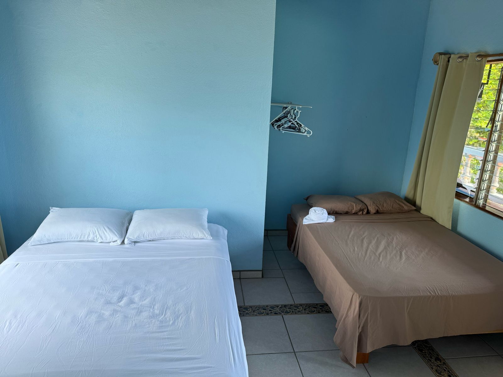 A room with two beds, one with white bedding and the other with brown bedding. There is a window with curtains, a wall-mounted drying rack, and a tiled floor.