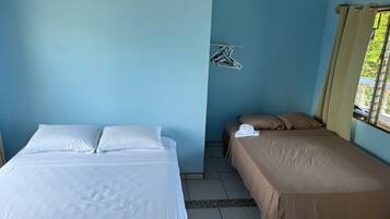 A room with two beds, one with white bedding and the other with brown bedding. There is a window with curtains, a wall-mounted drying rack, and a tiled floor.