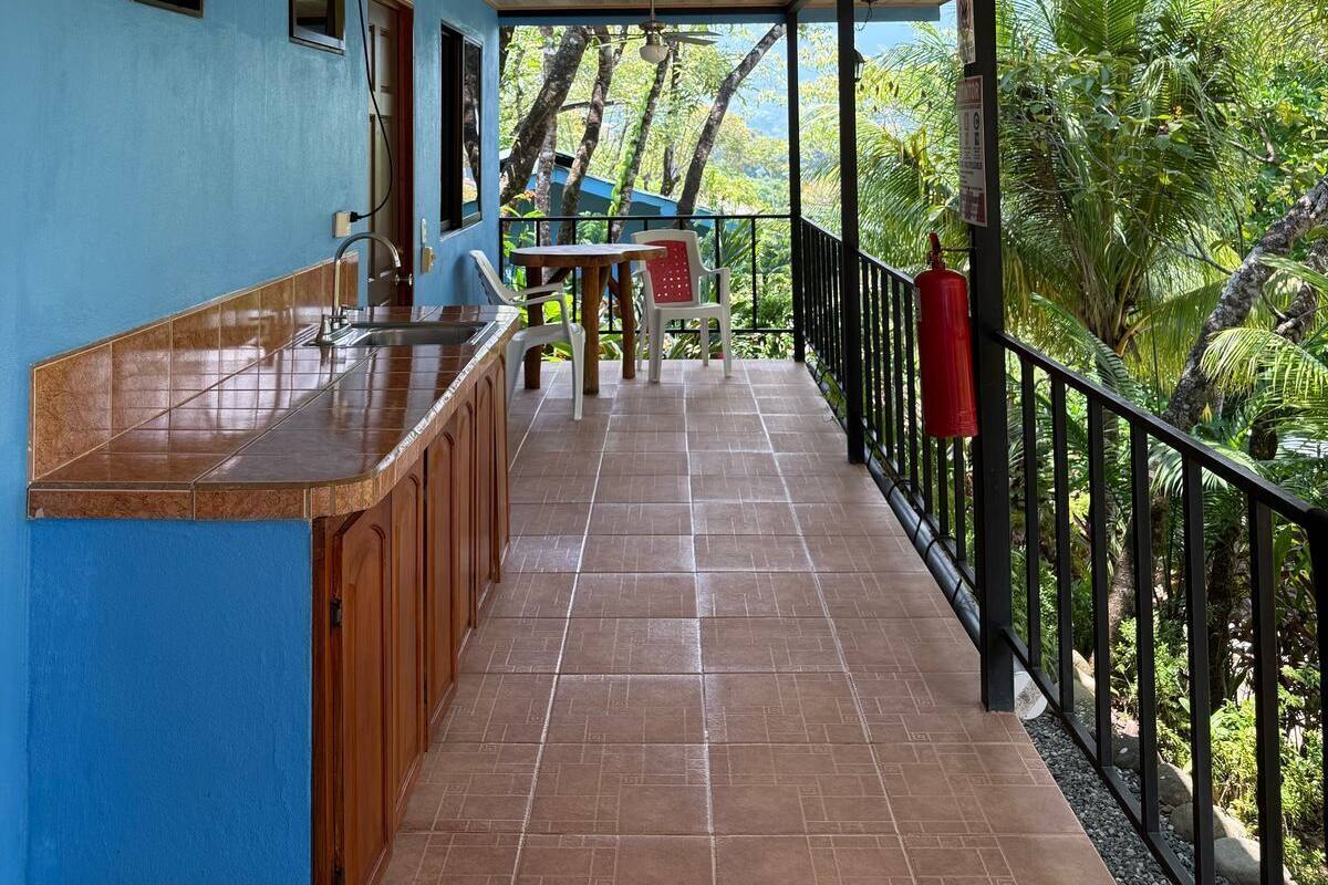 A covered outdoor area with a kitchen counter, a dining table, and chairs. There is a view of greenery through the railing.