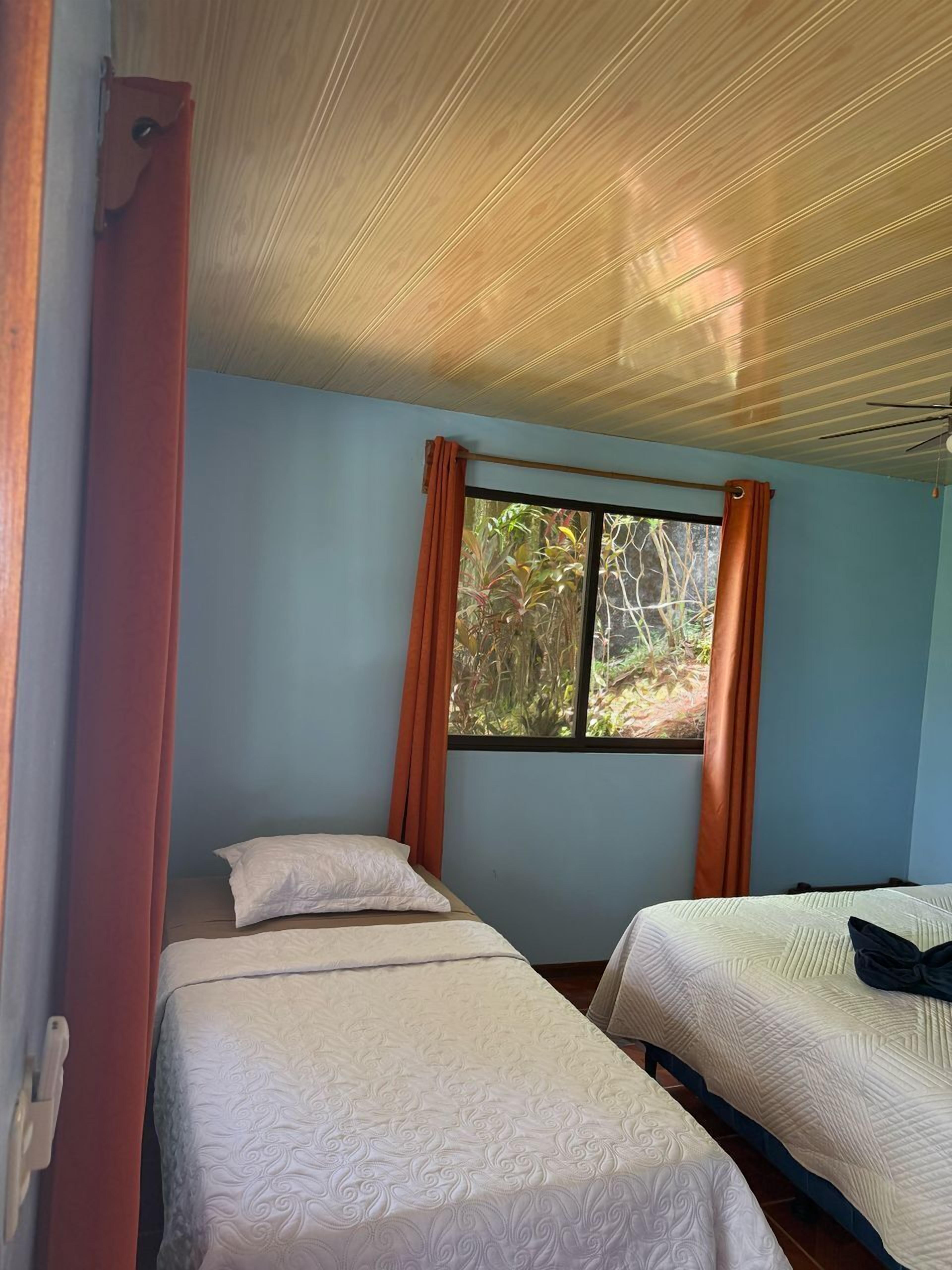 A room with two beds, a window with curtains, and a wooden ceiling.
