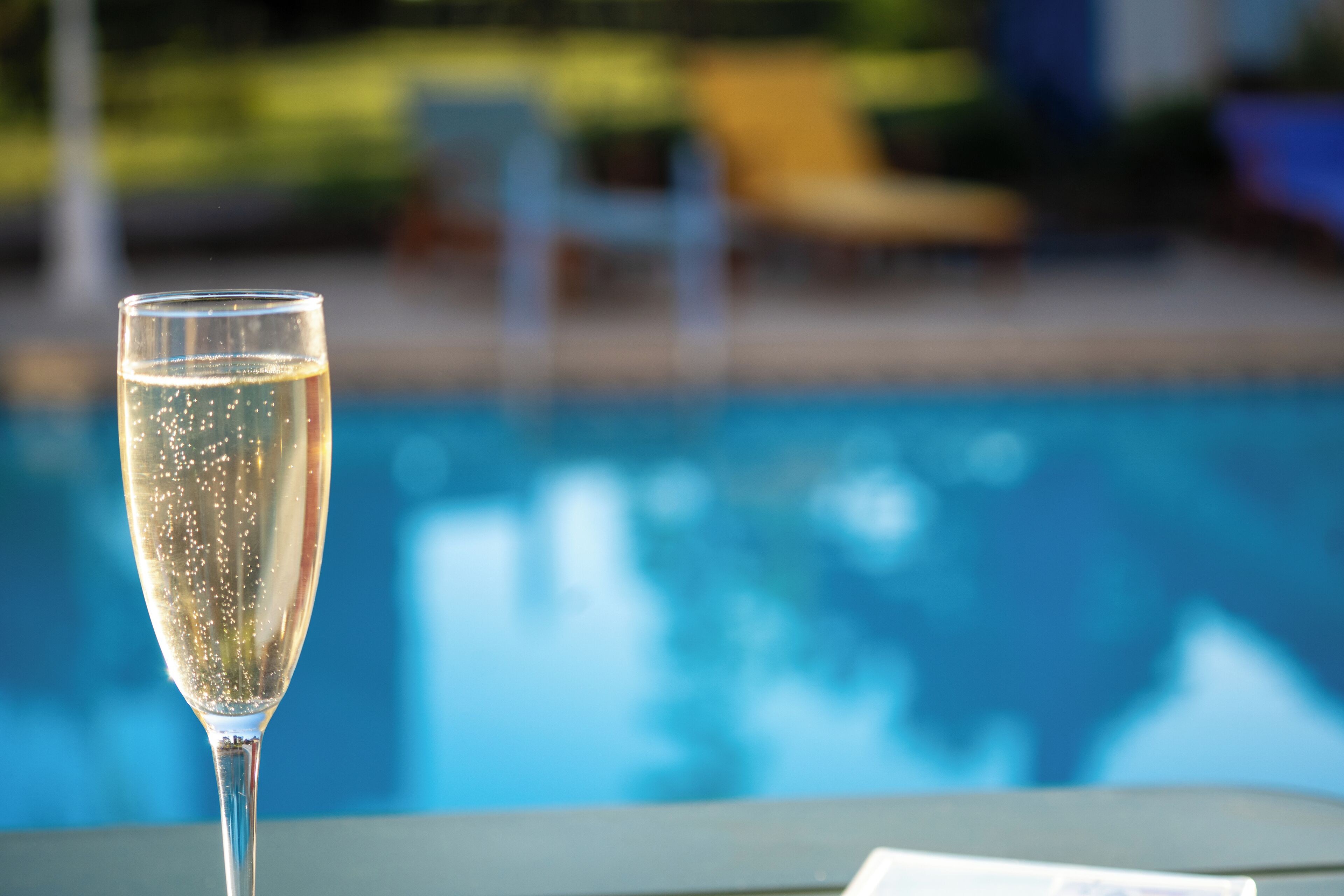 Un verre de champagne près d’une piscine, avec un arrière-plan flou représentant des tables et des chaises installées à l’extérieur.